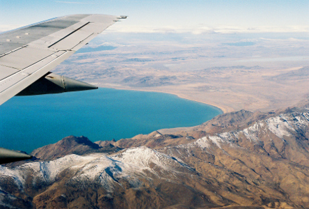 [The edge of the wing and two engines are visible along the left edge of the image. Partially snow-capped mountains are along the lower edge and partially up the right side. There is a very blue lake with what looks to be slightly low water levels under the wing and across the middle of the image. The upper part of the image is land on the same level as the lake. The ground is brown.]